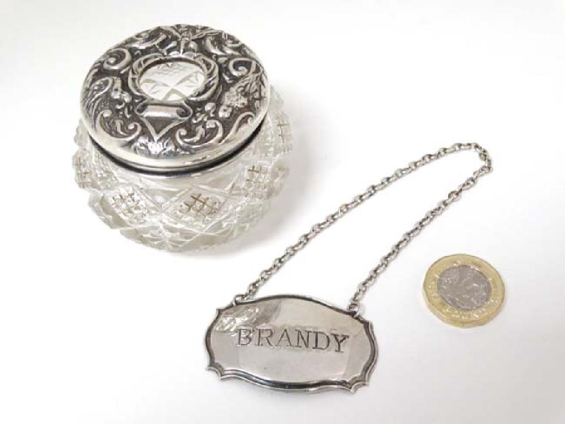 A cut glass hair tidy with silver top hallmarked: A cut glass hair tidy with silver top hallmarked Birmingham 1912 maker Sydney & Co. 2'' high. Together with a silver plate wine label ' Brandy ' (2)