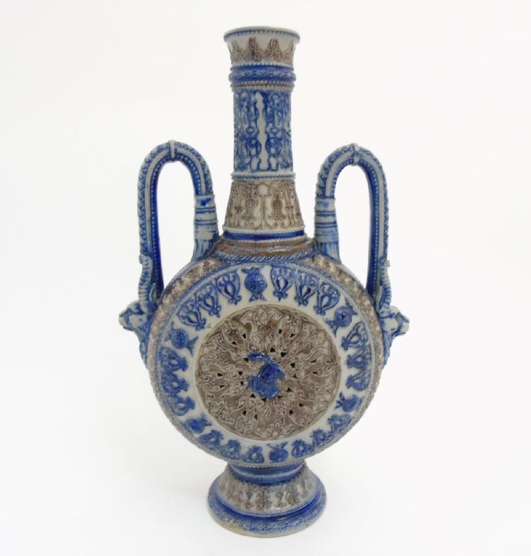 A Bavarian moon flask urn in the manner of Mettlach /: A Bavarian moon flask urn in the manner of Mettlach / Westerwald, having a circular body with tall flaring neck and handles having Capricorn heads, set on a pedestal foot base, surface decorated in re