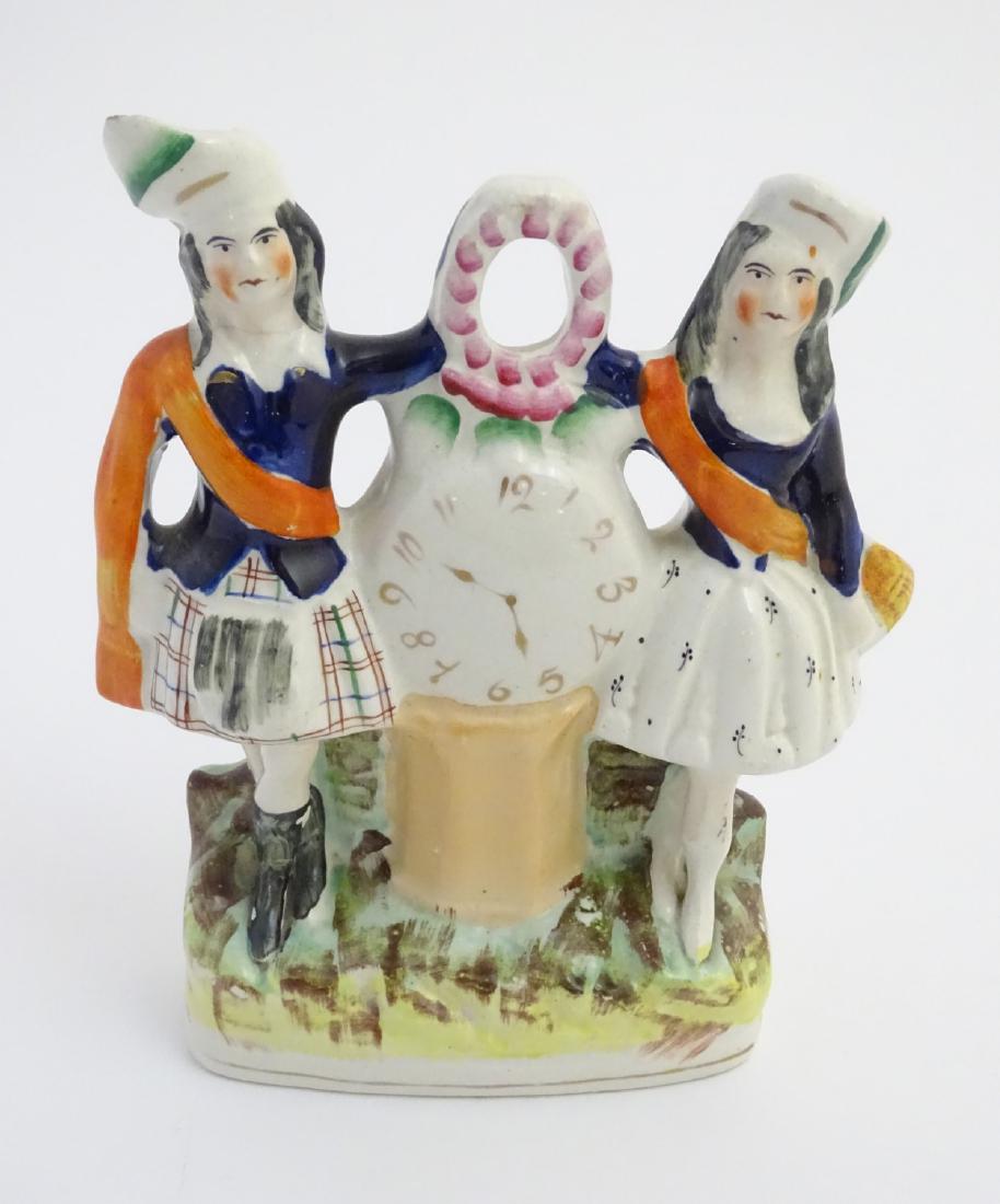 A Victorian Staffordshire pottery flatback figural: A Victorian Staffordshire pottery flatback figural group of a man and woman standing in highland dress either side of a clock face resting on a base with flowers above. Approx. 8 1/2'' high.