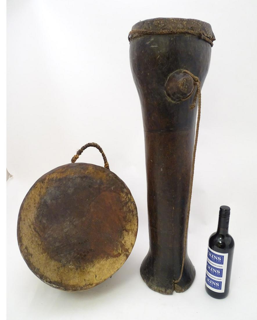 Ethnographic, Native, Tribal: two African drums one: Ethnographic, Native, Tribal: two African drums one tall with snake skin covering and plaited shoulder strap, together with a deer skin covered half sphere shaped drum with leather thong ties and carr