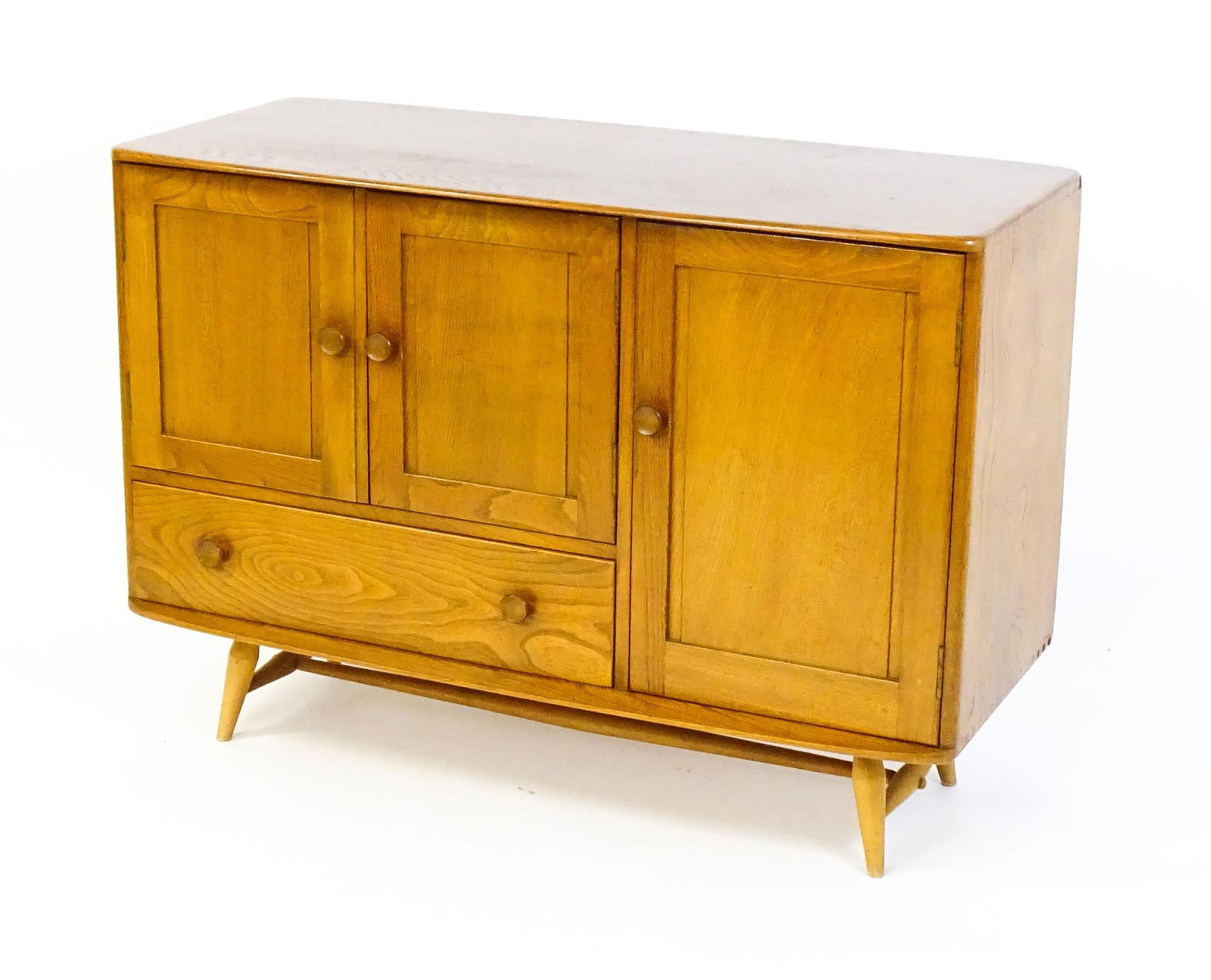 Vintage / Retro: An Ercol elm sideboard (model 366), the figured elm top above three panelled: Vintage / Retro: An Ercol elm sideboard (model 366), the figured elm top above three panelled cupboard doors and a long drawer, the superstructure raised on four turned tapering legs united by a H-str