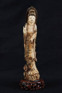 A RARE TINTED CARVED IVORY FIGURE OF GUANYIN: An ivory figure of Guanyin holding ruyi sceptre on the left while the other hand raised in a mudra of blessing, standing top of lotus sceptre, a shishi at her feet. Height: 10 1/2in (26.5cm)