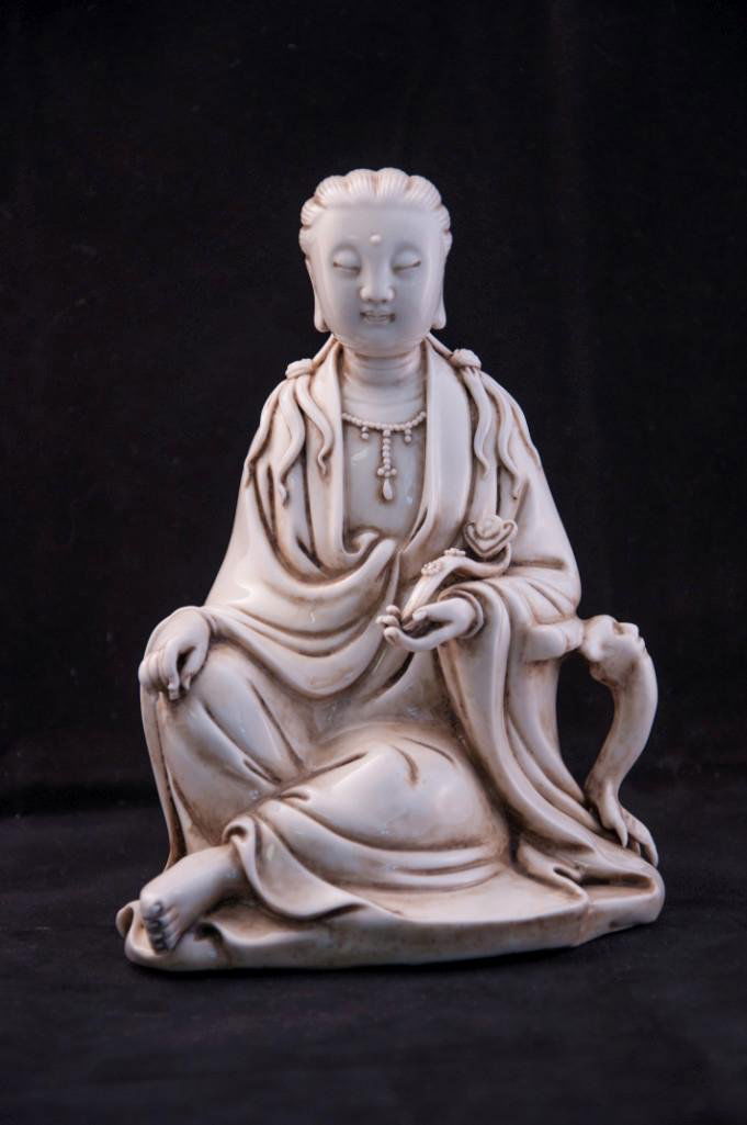 A CHINESE BLANC DE CHINE SEATED FIGURE OF GUANYIN: Modeled in a seated pose with flowing robes holding a Ruyi scepter in left and gracefully resting right hand, her hair dressed in knotted tresses, and her face modelled with gentle expression, all und