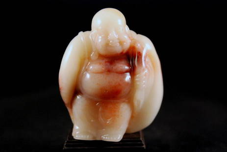 A CARVED SHOSHAN STONE BUDDHA WITH BINDLE: Of translucent pale gray-yellow and light orange stone with natural red-orange, carved figure of smiling Buddha with long ears leaning towards left with large sack. Height: 3in (7.5cm)