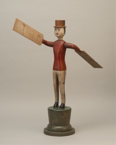 A Folk Art Whirligig: a standing man, wearing a top hat, painted red, white and black, standing on a marbled green plinth. 18 1/2" high.