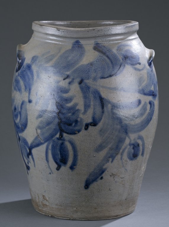 American Stoneware Crock: Two gallon stoneware jar with ovoid body, tooled and sloped rim and lug handles. Flowering vine wraps around the body and accents the handles. 13 3/4" H Condition: Two significant chips/losses to the