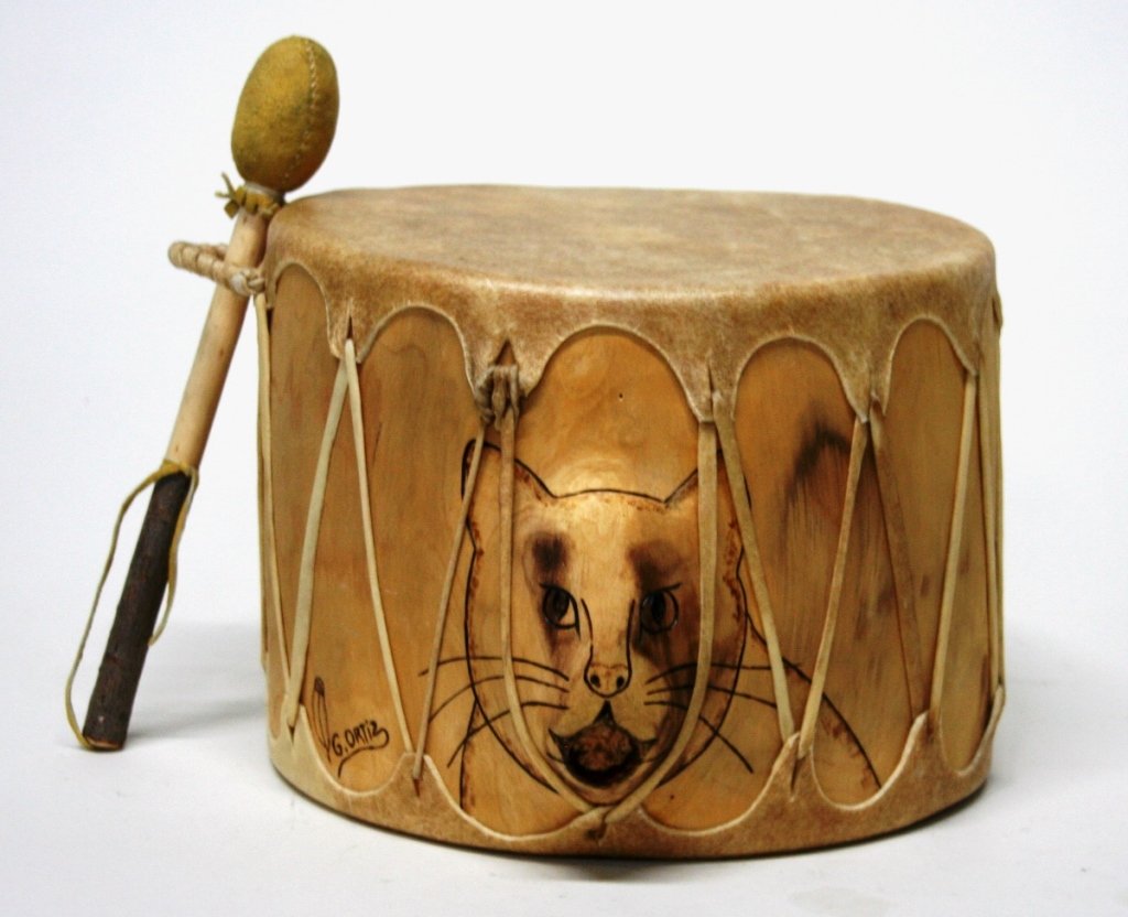 Cat Drum by G. Ortiz.: Wood, skin, and leather. Two high relief cat motifs, one on each side of drum. Signed to the left of the cat face "G. Ortiz" with a feather. 8 1/2"h x 11"diam.