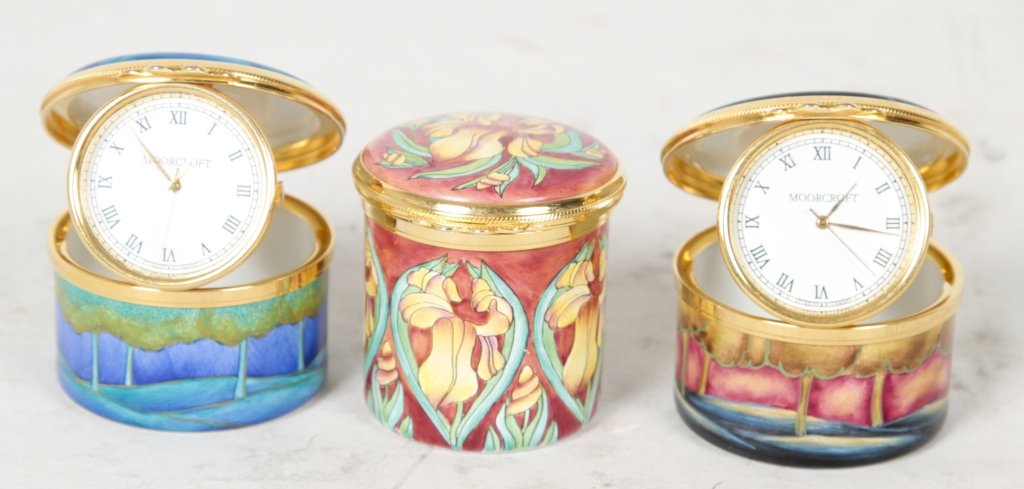 Three Moorcroft Enamel Pill Boxes: including: two which open to reveal small travel clock decorated in woode scenes, together with one in iris pattern. with clocks: H1 1/4" iris: H1 3/4" From the Estate of Anita Hill, Jupiter, Florida.