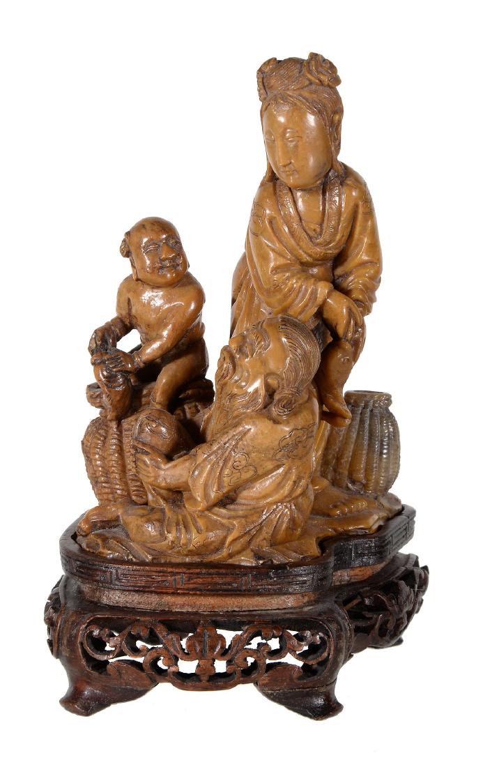 A Chinese soapstone group depicting an elderly: A Chinese soapstone group depicting an elderly fisherman seated, his wife beside him and a small boy seated atop a large fishing basket, with fitted wood stand, height overall 14.1cms; 19th-20th Centu