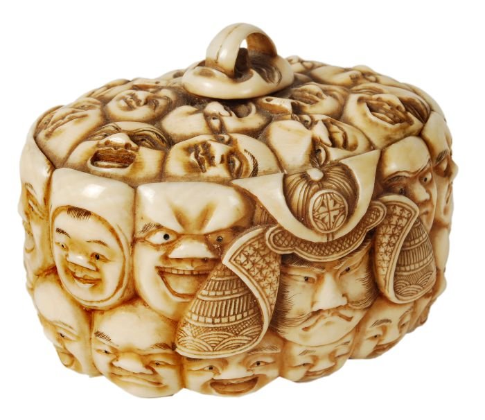 A Japanese ivory box and cover of oval form carved: A Japanese ivory box and cover of oval form carved all over with a profusion of comic masks, the knop of the cover formed from the protruding tongue of a mask, unsigned, length 7.4cm, Meiji period Pro