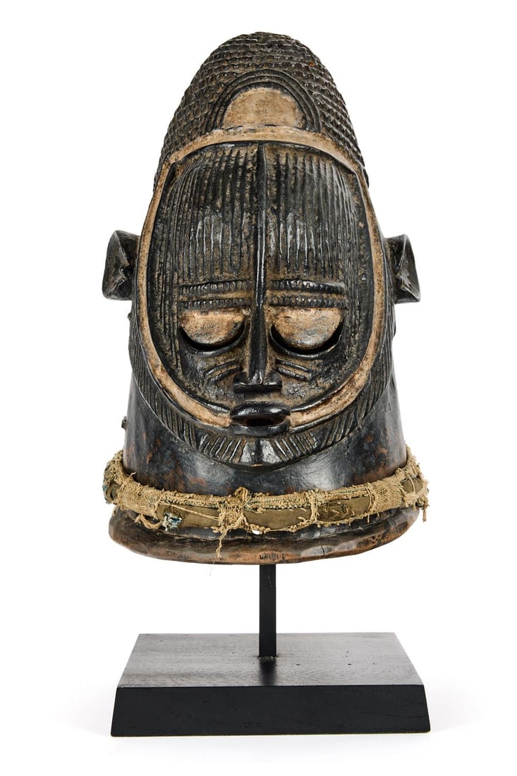 AN IGALA HELMET MASK: AN IGALA HELMET MASK NIGERIA, CIRCA 1910 The incised face with pointed chin, overall scarifications, all covered with black and white pigments, textile bundle around the neck, rich black thick patina