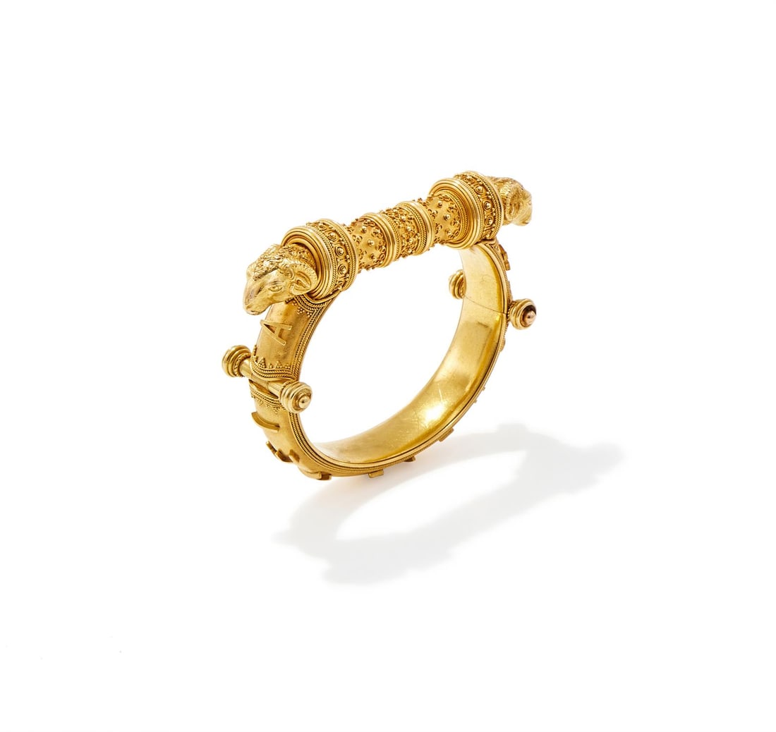 AN ARCHAEOLOGICAL REVIVAL BANGLE: AN ARCHAEOLOGICAL REVIVAL BANGLE1870SThe cylindrical bar with filigree and beadwork decoration, between ram's head finials, one a pencil holder, to stylized hinges with locking post clasp, applied scr