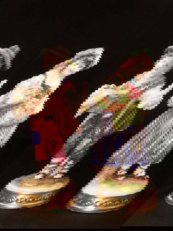 ANTIQUE VOLKSTEDT PORCELAIN FIGURES BOY GIRL: Both beautifully hand painted. Dressed in peasant style tattered clothes. Girl holds a basket of vegetables and boy is holding a figurine pulled from a basket. Both stand on round bases trimmed hand p