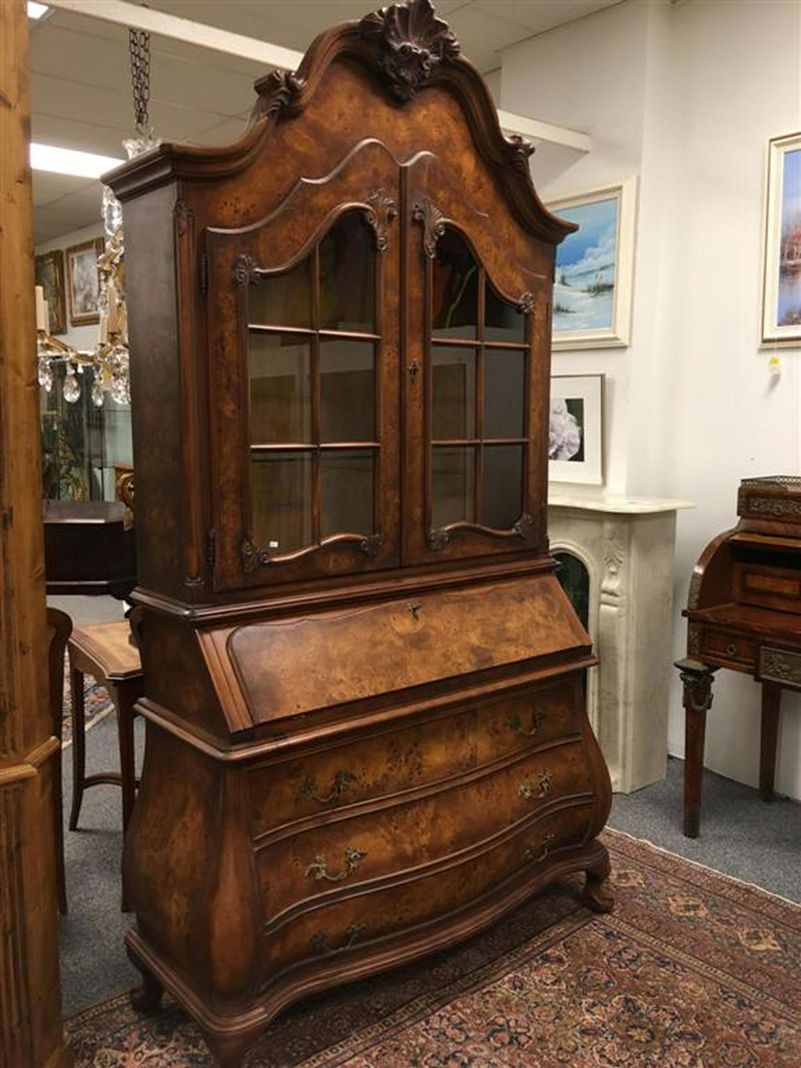 A BURLED AND CARVED WOOD SECRETARY: A burled and hand carved secretary. Two locking glass doors, two adjustable glass shelves, illuminated interior, locking drop lid with pill-out supports, fitted interior with four drawers, bombe style