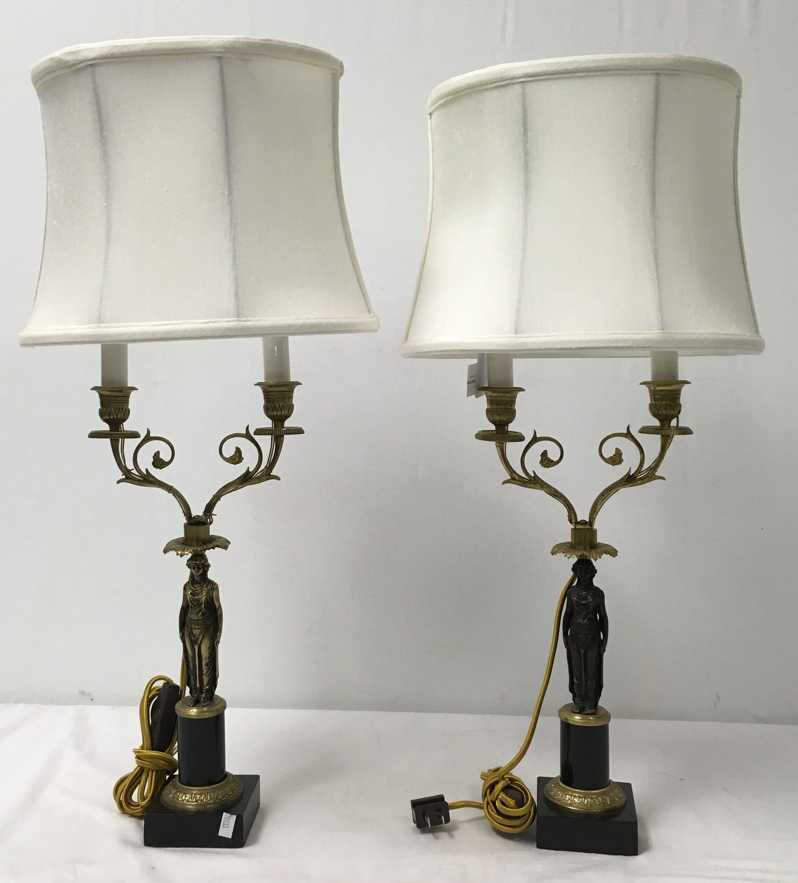 PAIR NEOCLASSICAL DOUBLE LIGHT CANDELABRA AS LAMPS: Pair of Neoclassical double light candelabra, mounted as table lamps. Each one includes an off-white fabric shade and measures a total of 25.75" tall x 12" wide (shade width). Base width is 3.25", wid