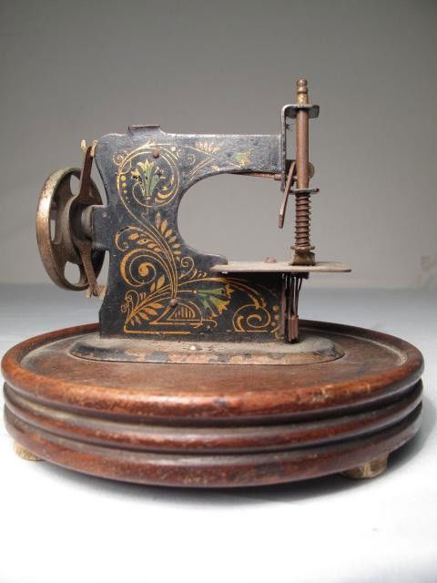 MINIATURE ANTIQUE GERMAN SEWING MACHINE: Miniature antique German hand crank sewing machine on round wood stand. Black painted with stencil design. Stamped Made in Germany. Some rust and wear. Gears turn smoothly. Measures 5" long by 2" wide