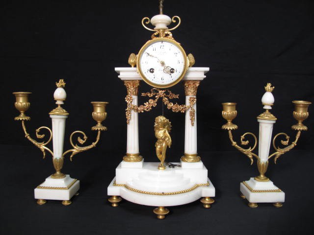 FRENCH CARVED MARBLE MANTLE CLOCK & CANDELABRAS: French carved marble mantle clock and candelabras. Ormolu mounts and hardware on white marble. Clock has a cherub figurine in front of sun-face motif pendulum. Pillars holding up the clock above a sma
