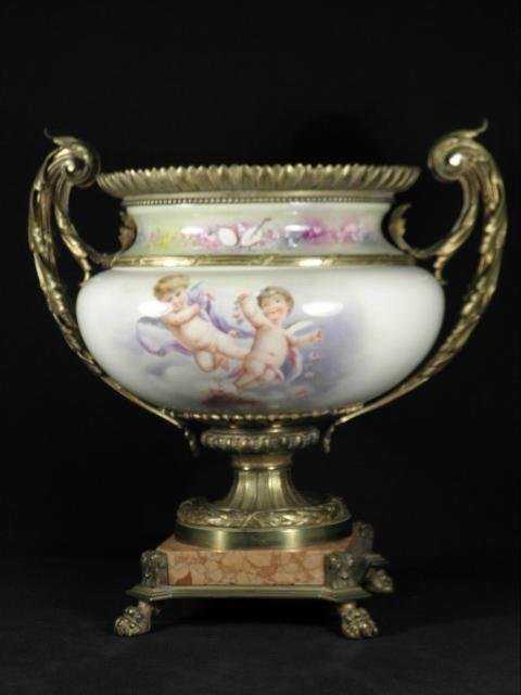 BRONZE MOUNTED HAND PAINTED PORCELAIN URN: Bronze mounted hand painted porcelain urn. Ormolu gilt bronze handles and gallery around rim, with a cut marble base, all on four claw feet. Hand painted with a cherub scene and highlights with floral