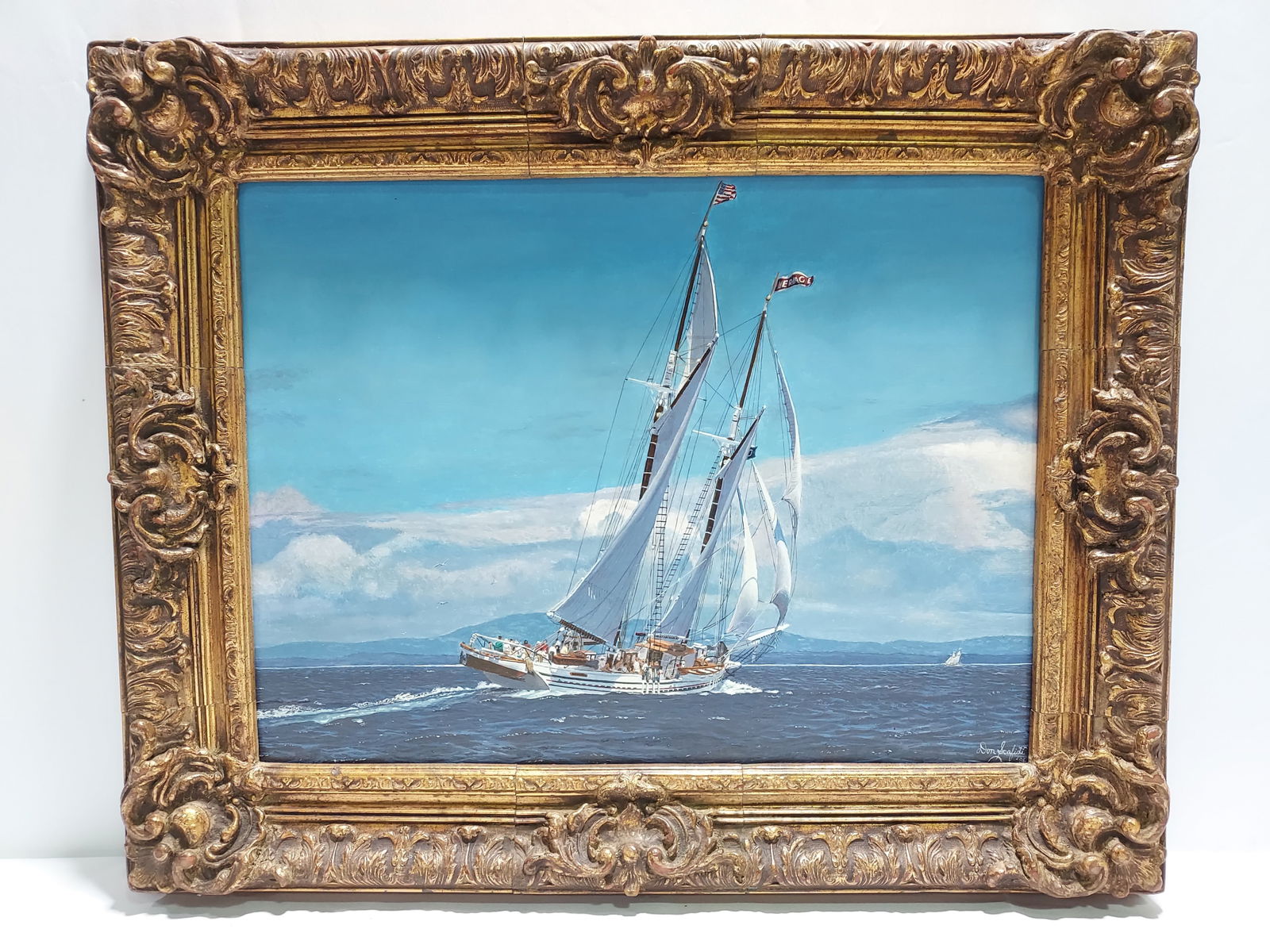 DON SCAFIDI S/V HERITAGE OIL ON BOARD PAINTING: Don Scafidi oil on board painting of the S/V Heritage. Painting depicts a large sailboat with passengers, a mountainous background and a smaller sailboat in the distance. Framed in an intricate gold t