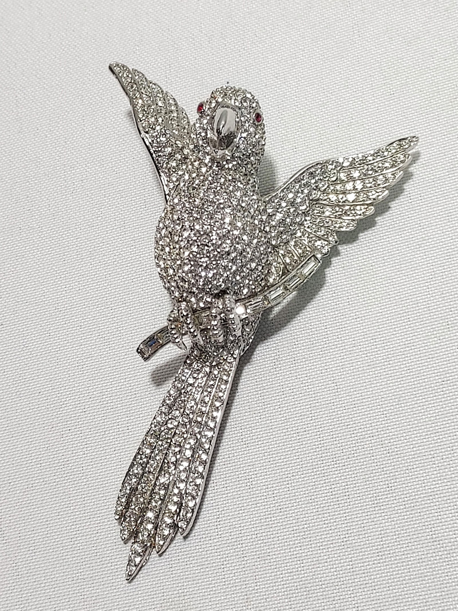 H607 Ciner bird brooch: Ciner bird brooch: silver tone Ciner rhinestone encrusted bird on a branch brooch. Bird has red rhinestone eyes and is perched on a baguette cut rhinestone branch