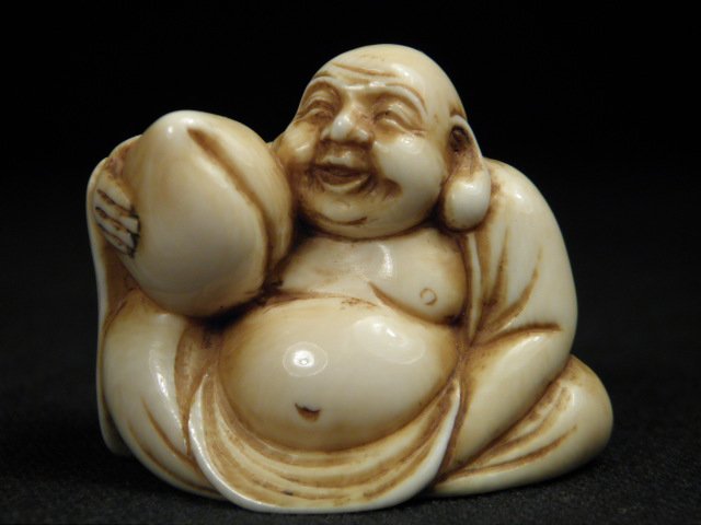 JAPANESE CARVED IVORY NETSUKE: HOTEI WITH PEACH: Japanese carved ivory netsuke depicting a laughing Hotei figure holding a peach. Signed with artist's signature on the base near loop holes. Condition: very minor discoloration as expected from age, s