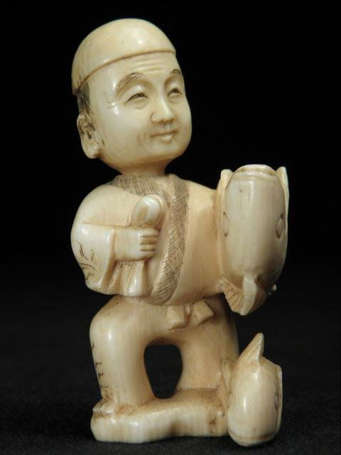 JAPANESE CARVED IVORY NETSUKE: BOBBLE HEAD FISHERMAN: Japanese carved ivory Karakuri netsuke depicting a fisherman holding a fish. Bobble head design, with darkened details and artist's signature on base. Condition: small age crack near loop hole, minor