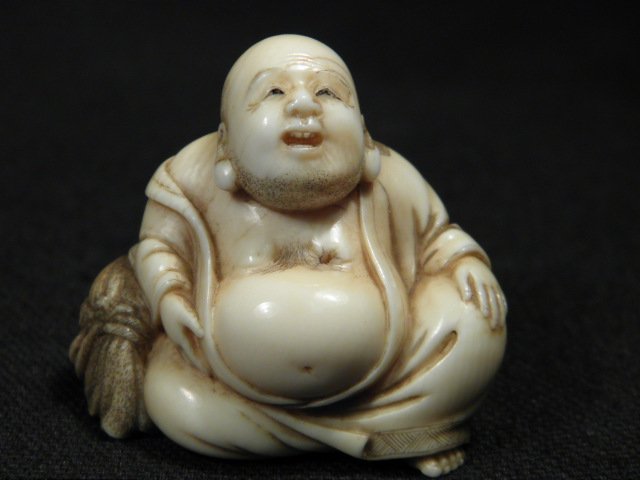 JAPANESE CARVED IVORY NETSUKE: SEATED HOTEI: Japanese carved ivory Katabori netsuke depicting a seated Hotei looking upwards. Black highlighted in the details, signed on base, and with loop holes on the backside. Condition: small hole to ivory o