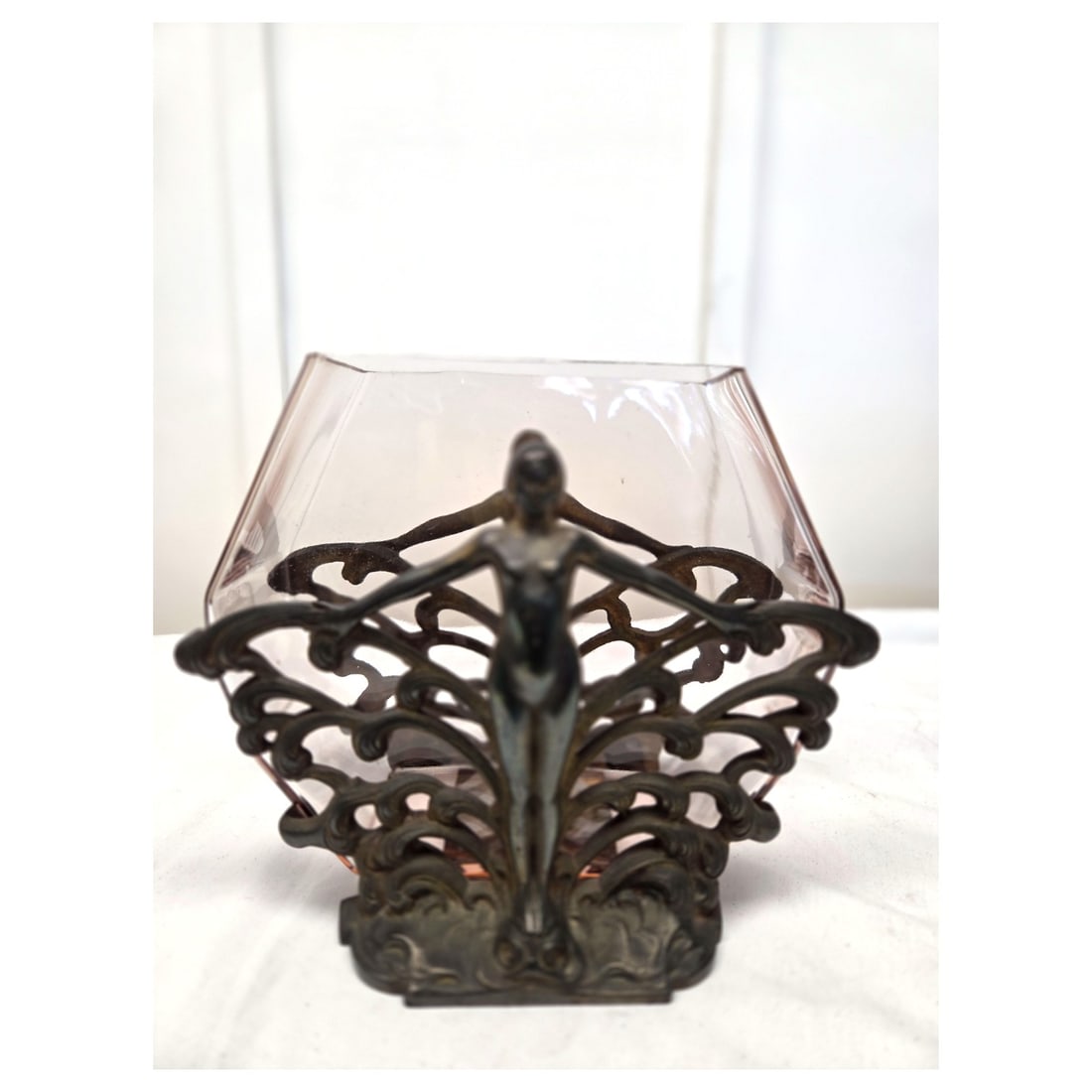 ART DECO IRON AND PINK GLASS TABLETOP FISH BOWL: An Art Deco fish bowl. The cast metal fish bowl stand has female figures with a wave motif on each side. It holds a hexagonal light pink glass fish bowl. Measures: Overall 9.5" T x 11" W x 7.5" D.