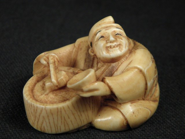 JAPANESE CARVED IVORY NETSUKE: MAN WITH BOWL: Japanese carved ivory netsuke depicting a sitting man serving food from a bowl. Signed by artist on base (near loop holes). Condition: very minor discoloration as expected, see images. Measures 1.1" t