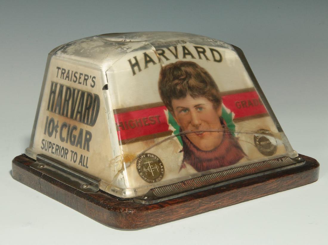 HARVARD BRAND CIGARS ADVERTISING CIGAR CUTTER: A glass counter-top cigar cutter with damage, the thick glass housing mounted to oak base contains the key-wind clockwork cutting mechanism triggered by inserting the cigar tip into the blade area on 