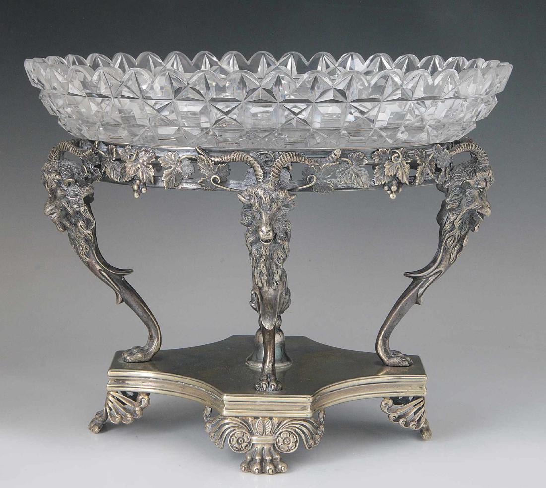 A SILVER PLATE CENTERPIECE WITH CUT CRYSTAL: Quadripartite stand raised on four well-defined paw feet headed by stylized scrolls and rosettes supporting four horned goat head monopodiae terminating in paw foot, the heads linked by a pierced Bacc