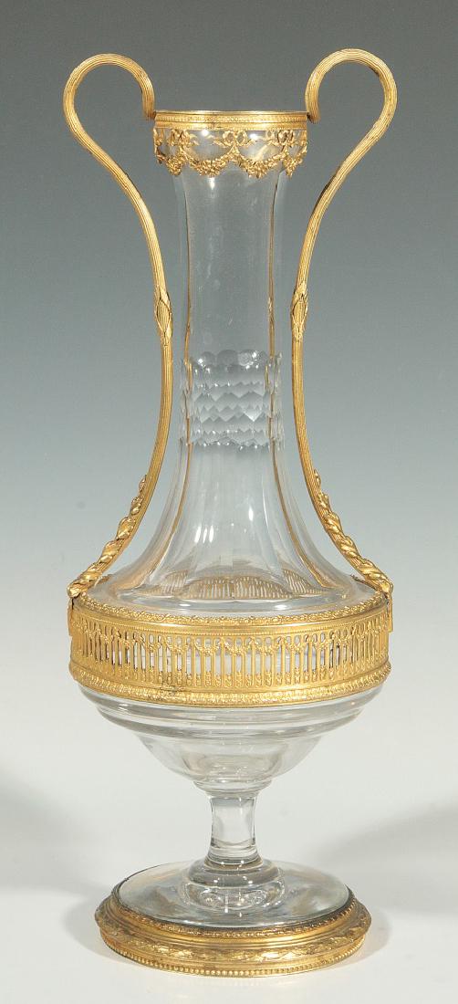 AN ORMOLU MOUNTED LOUTROPHOROS FORM CRYSTAL VASE: An early 20th century Loutrophoros-form vase with gilt overlay openwork band to short body, the elongated neck with cut panels flanked by long graceful reeded handles draped with leafy motif connectin