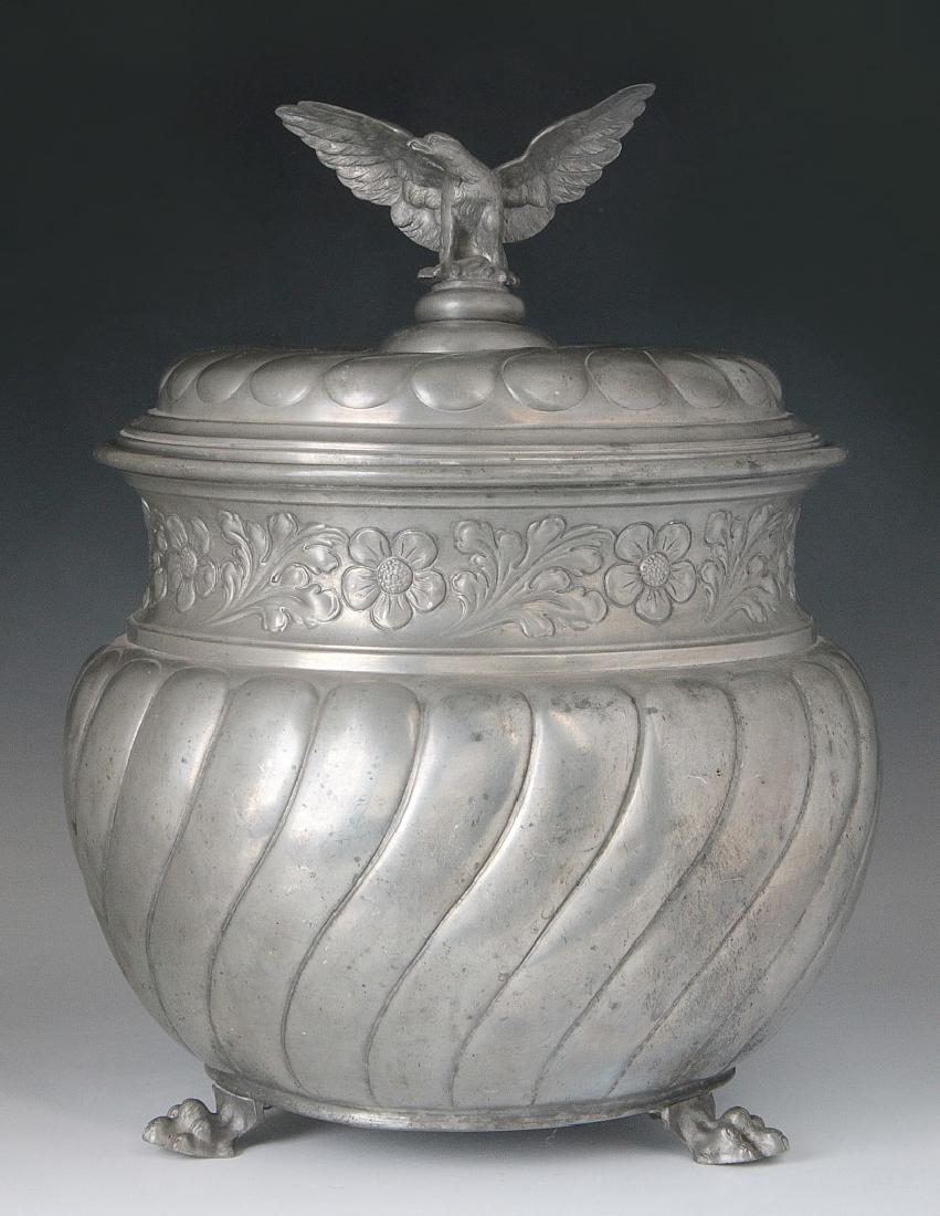 A 19TH CENTURY PEWTER WINE COOLER: Bulbous twisted lobed body with tapering sides having waisted neck decorated with a band of flowers and foliate in relief, the low domed lid of conforming lobed design and surmounted by an eagle finia