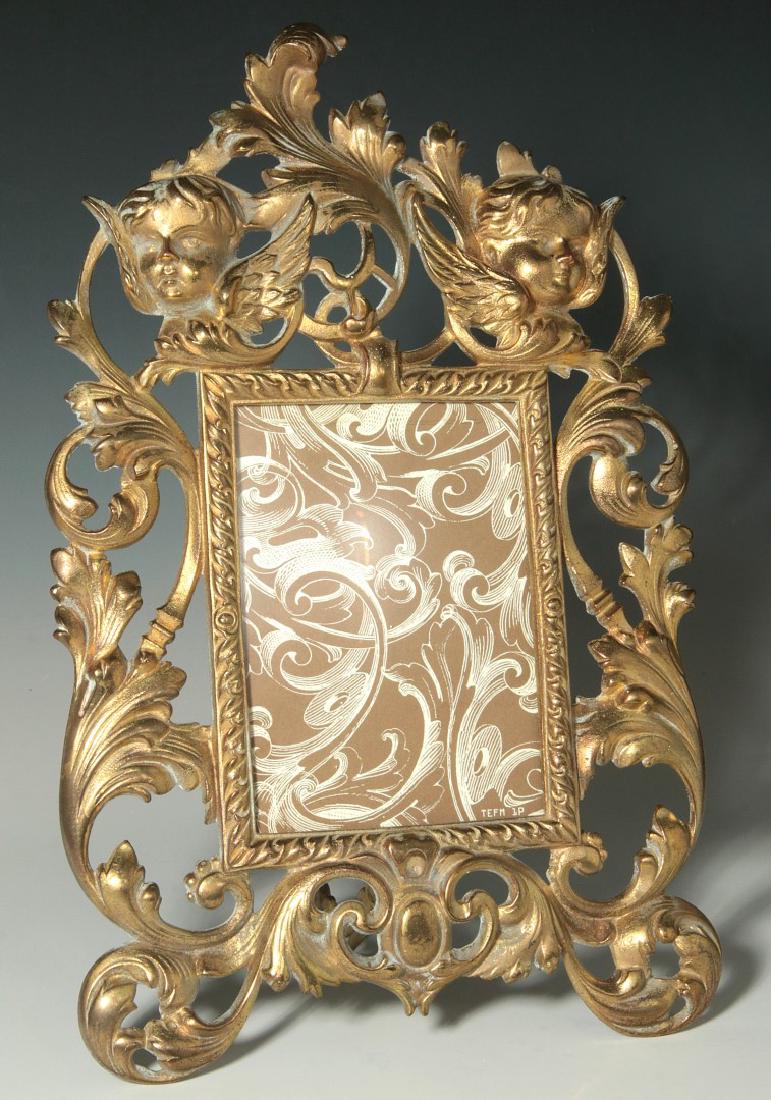 A ROCOCO STYLE BRONZE EASEL BACK FRAME WITH PUTTI: Florentine Gilt Bronze Frame, The rectangular frame within a pierced scrolling acanthus border, surmounted by a crest of two putti masks which flank an acanthus leaf, the bottom centered by a cabochon