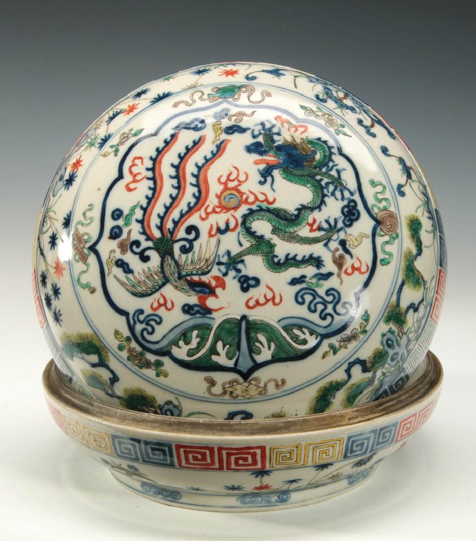 A LARGE AND EARLY WUCAI PORCELAIN COVERED BOX: The lid decorated with design of a five claw dragon and phoenix over waves within a ruyi shaped cartouche, the sides decorated with Buddhist symbols over branches and garden rocks and a key pattern ba