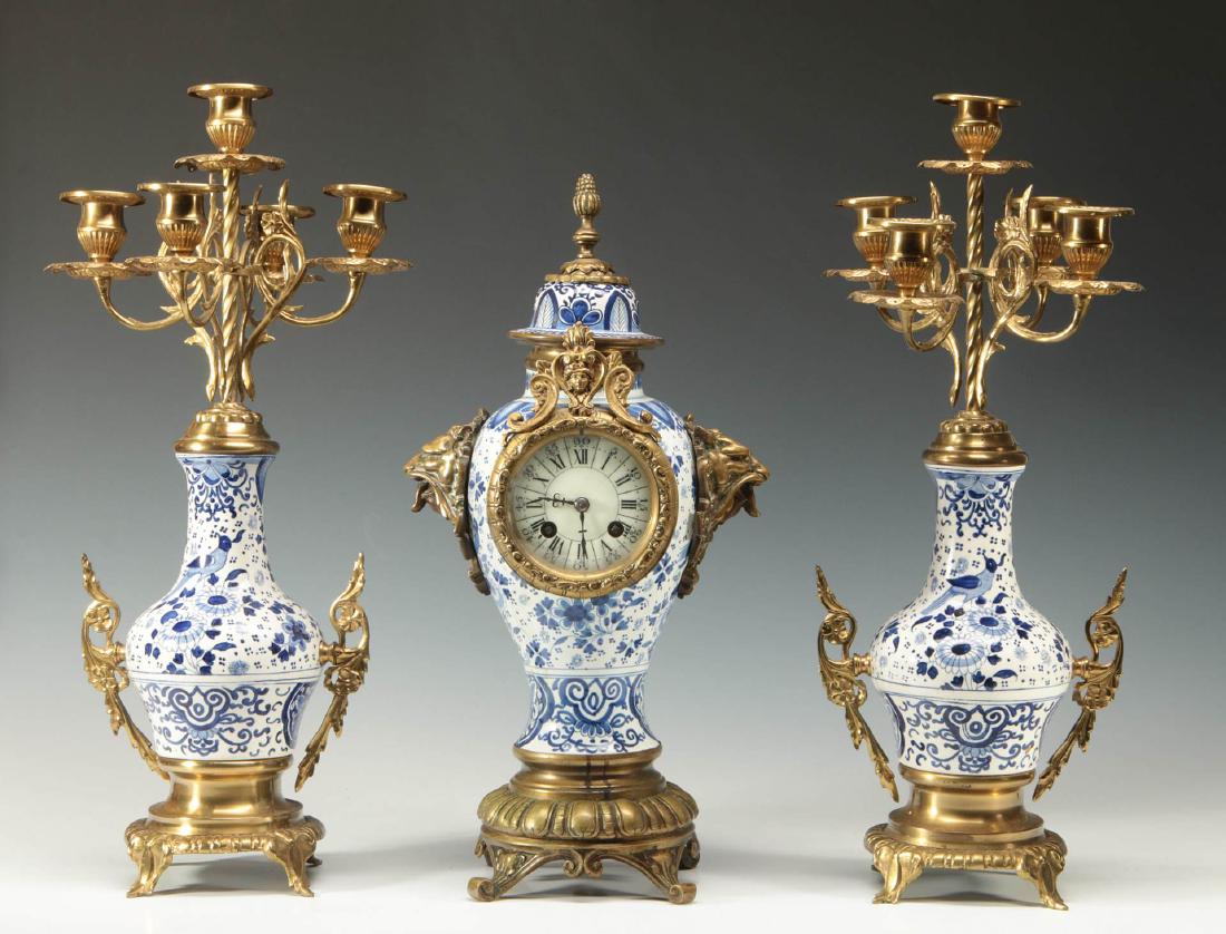 A BRONZE MOUNTED CONTINENTAL PORCELAIN CLOCK SET: A three-piece clock garniture, blue and white porcelain mounted with brass or bronze, the clock topped by an acanthus finial on high dome cover bolted to the baluster form body with lion masks either 