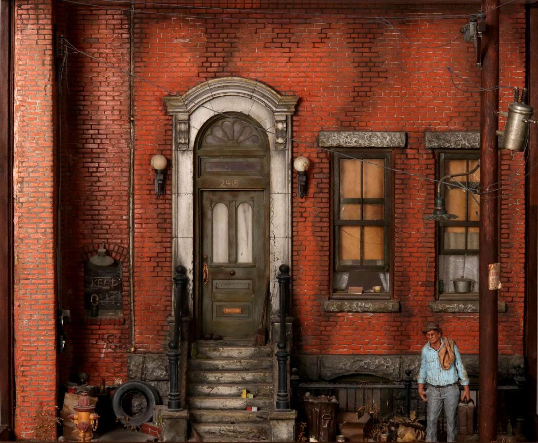 A MICHAEL GARMIN (B. 1938) CITYSCAPE DIORAMA: A gritty three-dimensional brownstone entrance; a figure in blue shirt carrying a suitcase outside the brick residence with stoop leading to a city sidewalk filled with everyday objects in great detai