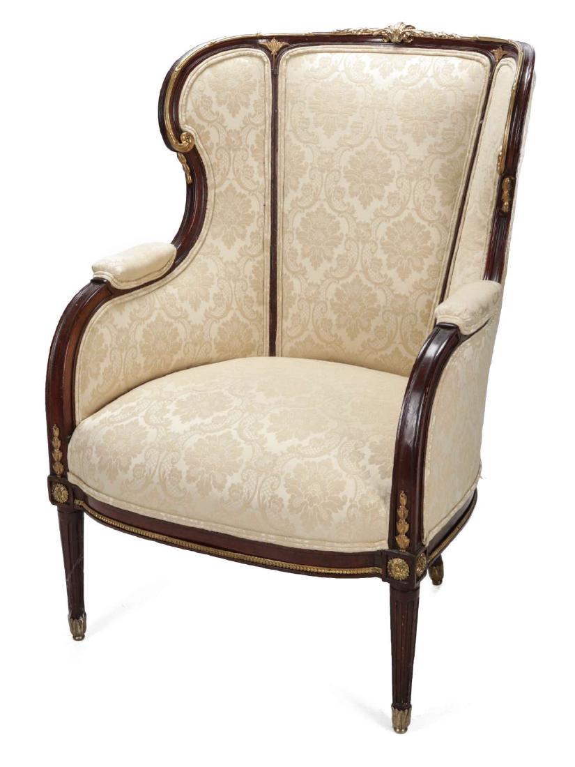 AN EARLY 20TH C. LOUIS XVI STYLE MAHOGANY BERGERE: The molded crest centering a gilt pendant flanked by berried laurel continuing down to padded armrests punctuated by gilt foliate and bellflower mounts, a tripartite padded back and sides over a tight