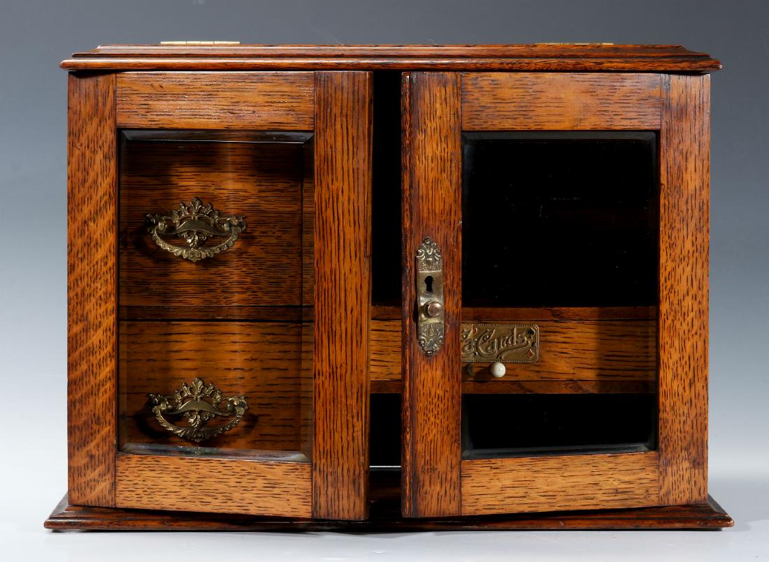 A CIRCA 1920s OAK PLAYING CARD AND GAMES CABINET: A good table-top oak cabinet with hinged front doors set with beveled glass, the fitted interior divided with two drawers and a more narrow drawer to the side with an embossed and enameled brass tag l