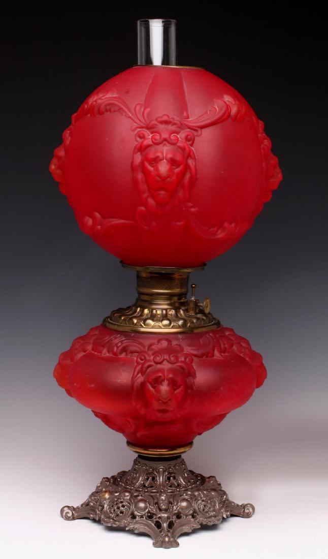 A FROSTED BLOWN-OUT CRANBERRY GLASS PARLOR LAMP: The upper ball-shape shade with blown out design of lion's heads and scrolls in cranberry satin glass, brass burner and font on matching glass base mounted to a rococo style spelter base, circa 1880. 