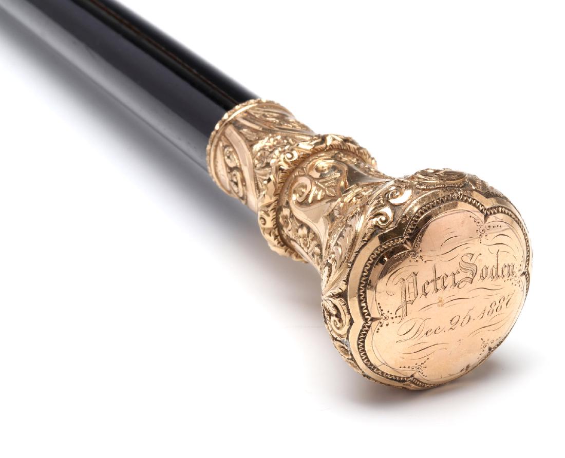 A FINE GOLD HANDLE WALKING STICK ENGRAVED 1887: The tapering ebonized wood stick with brass ferrule topped by an elaborately chased and hand engraved knob handle, the gold or gold-filled metalwork rich in deeply-cut scrolls, leaves, flower heads an