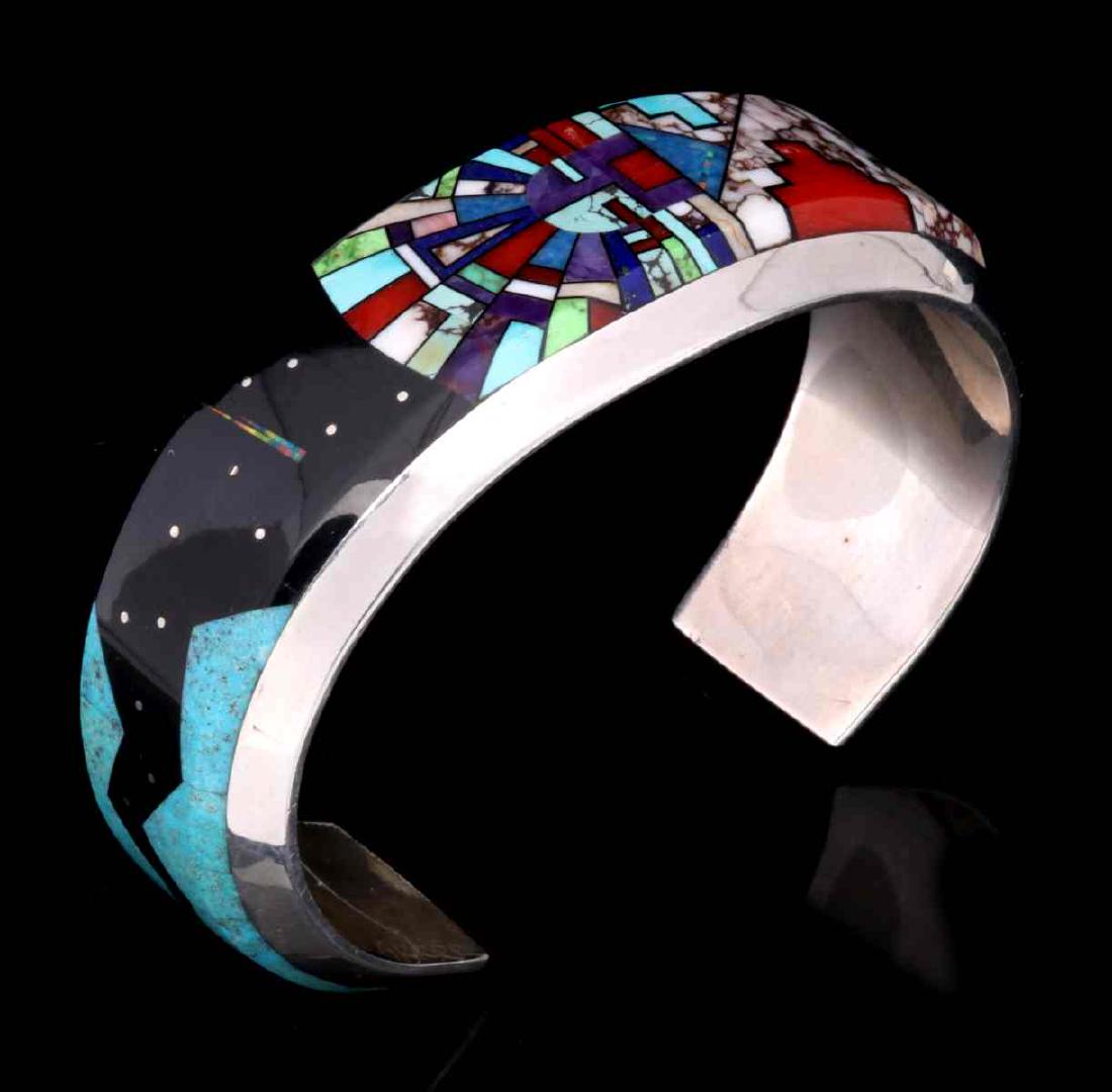 AN INLAID ZUNI STORYTELLER CUFF SIGNED SCOTT DAVE: The intricate inlay depicting five cultural themes: a highway running through Monument Valley, Kachina mask and headdress, a comet shooting through the night sky, a pueblo at night, and a spirit line 