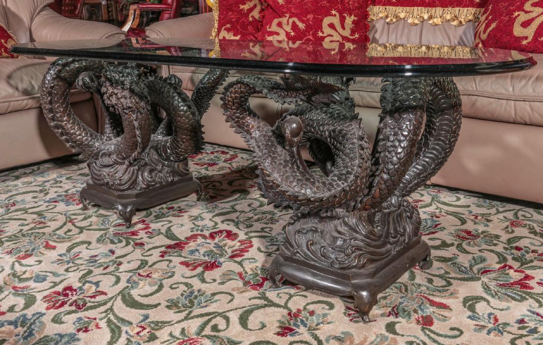 LONG OVAL COFFEE TABLE WITH BRONZE DRAGON SUPPORTS: The thick oval plate glass top with coved edge supported by two highly detailed cast bronze Chinese dragons clutching flaming pearls. Measures 20.5 x 60 x 24 inches. Provenance: Spirit of Avalon Mansi