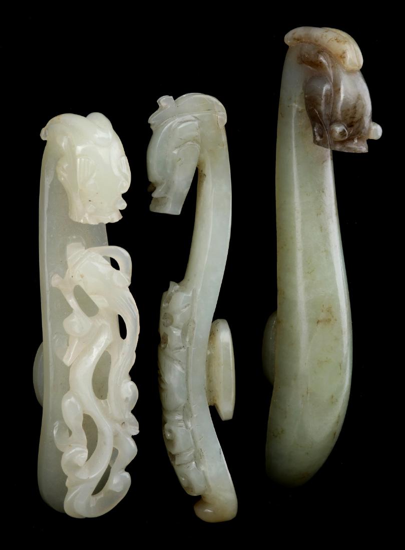 THREE CHINESE CARVED JADE DRAGON FORM BUCKLES: White and green jade ornamental clothing buckles, two with openwork carving, each with dragon head and 'button', 20th century. Range in length from 3.5 to 4.25 inches. We happily provide seamless in-h