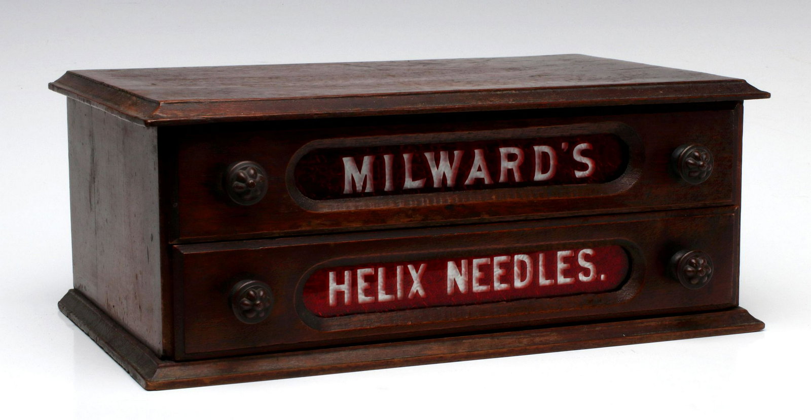A TWO-DRAWER MILWARD'S NEEDLES STORE CABINET: The walnut case with two drawers, each fronted by red-cut-to-clear glass inserts lettered with the maker's name and product, original fancy embossed brass knobs, intact divided top drawer, circa 1880.