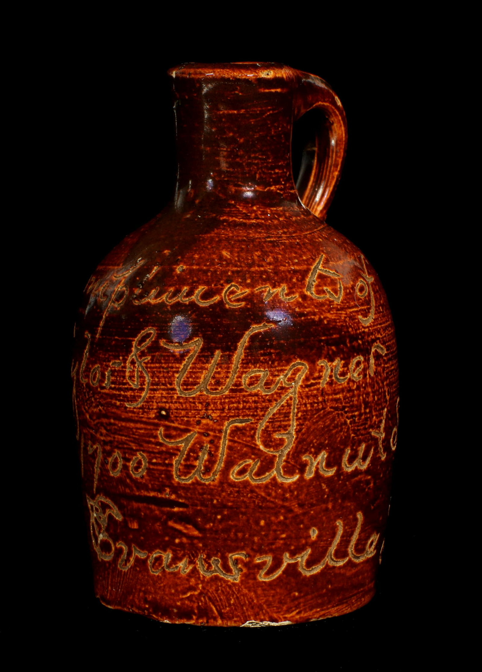 AN EVANSVILLE INDIANA SCRATCH STONEWARE MINI JUG: The Albany slip glaze etched 'Compliments of Taylor and Wagner 1700 Walnut St. Evansville, Ind.' Measures 3.5 x 2 inches. Provenance: The 45-year collection of Michael and Donna Hayes, West Plains, Mi