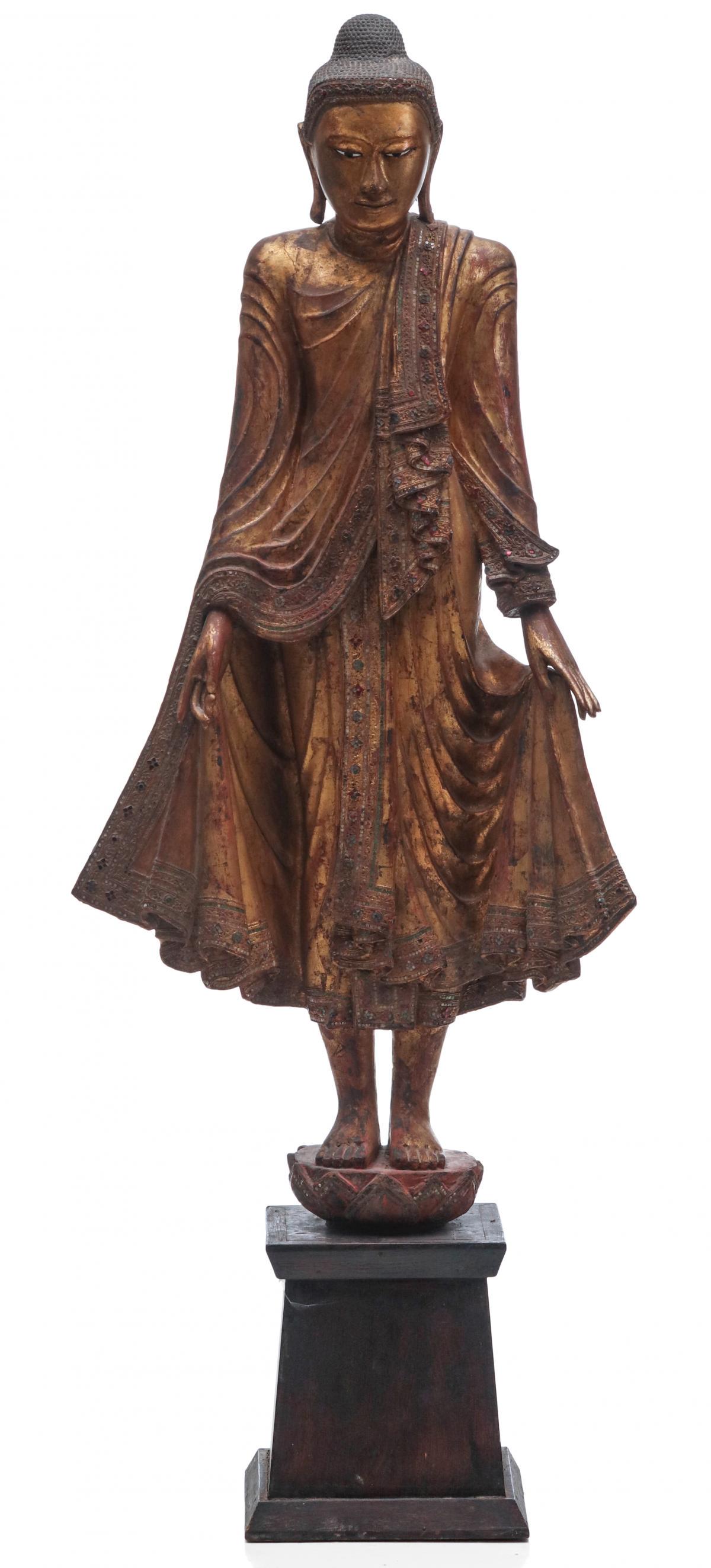 A 61-INCH CARVED AND GILDED WOOD THAI FIGURE OF BUDDHA: The carved and gilded wood full length figure standing upon on lotus and dressed in flowing robes with inlaid glass jewels, mounted, on an ebonised wood plinth. Measures 61 x 23 x 11.5 inches. We happ
