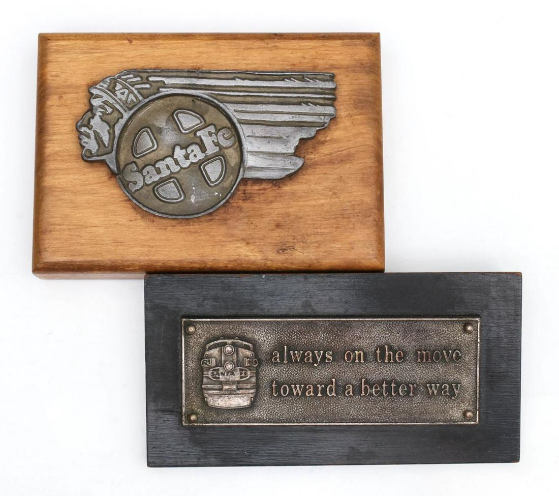 TWO SMALL MOUNTED SANTA FE RAILROAD BADGES: One paperweight pictures the front view of a Santa Fe Diesel engine 'always on the move toward a better way'; plus a chief logo badge mounted to wood block. Measures 2.5 x 5 x 0.75 and 3.25 x 4.75 x 1