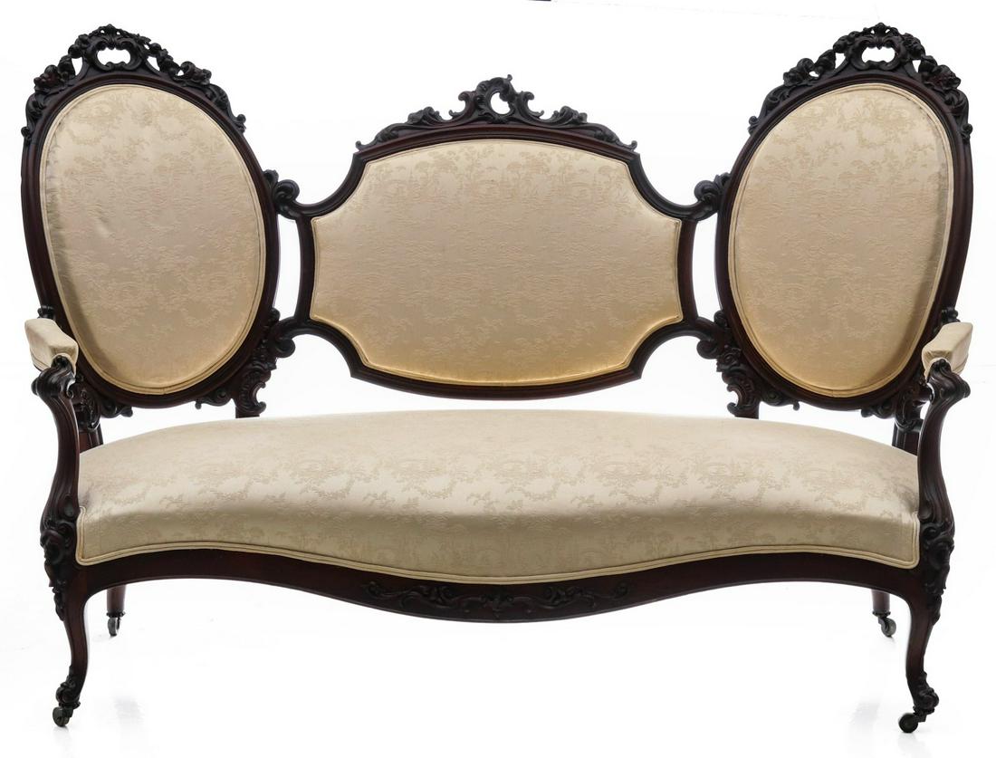 A 19TH CENTURY AMERICAN ROCOCO REVIVAL CARVED SOFA: The back comprised of two oval medallions and a central shield carved from walnut in Rococo taste and upholstered in an ivory damask, the overstuffed seat with open arms, the whole raised on short cab