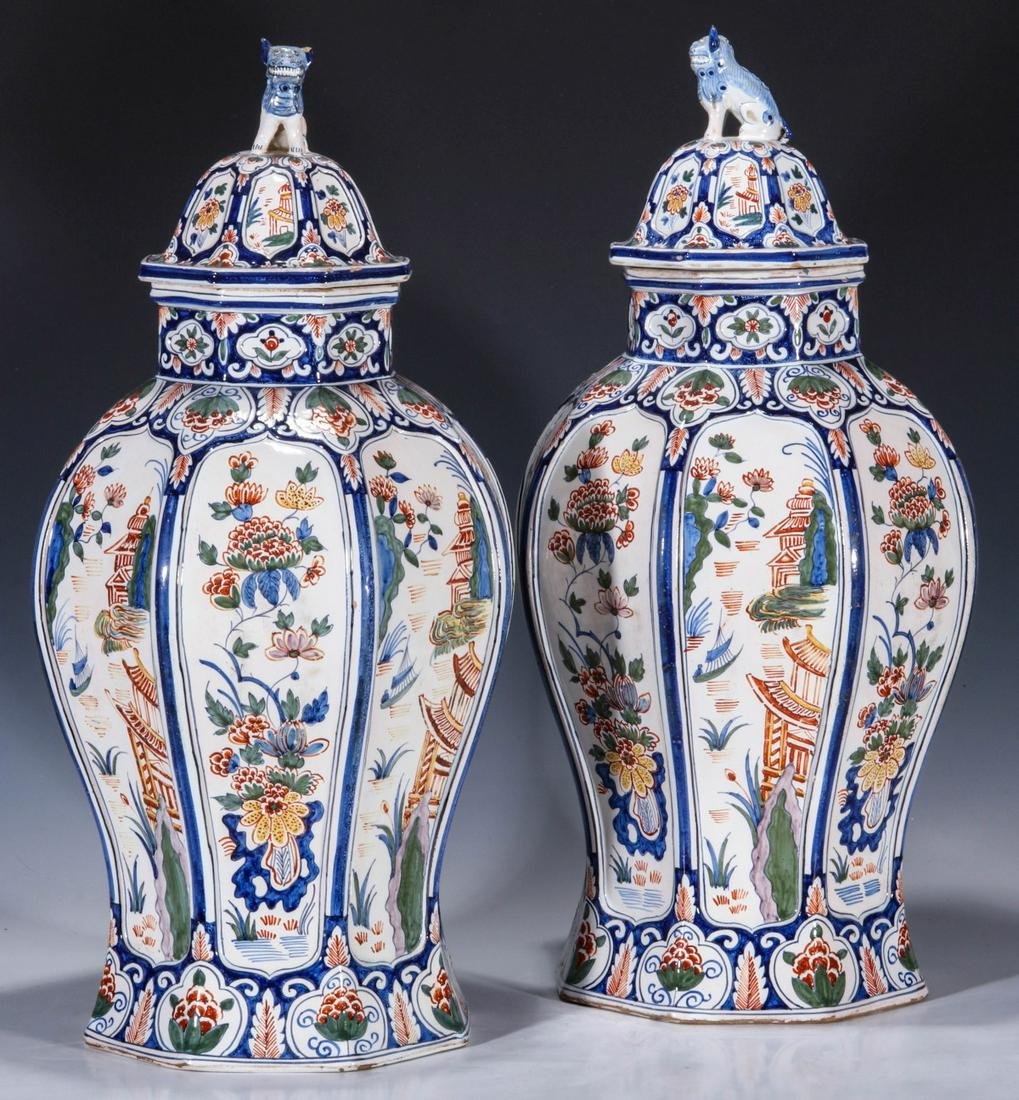 A PAIR 23-INCH LAMPETKAN DELFT POTTERY COVERED VASES: Each eight-sided vase with cover and alternating panels decorated in the Chinese taste with hand painted architectural and floral motifs, each cover mounted with foo dog finial, signed underside of th
